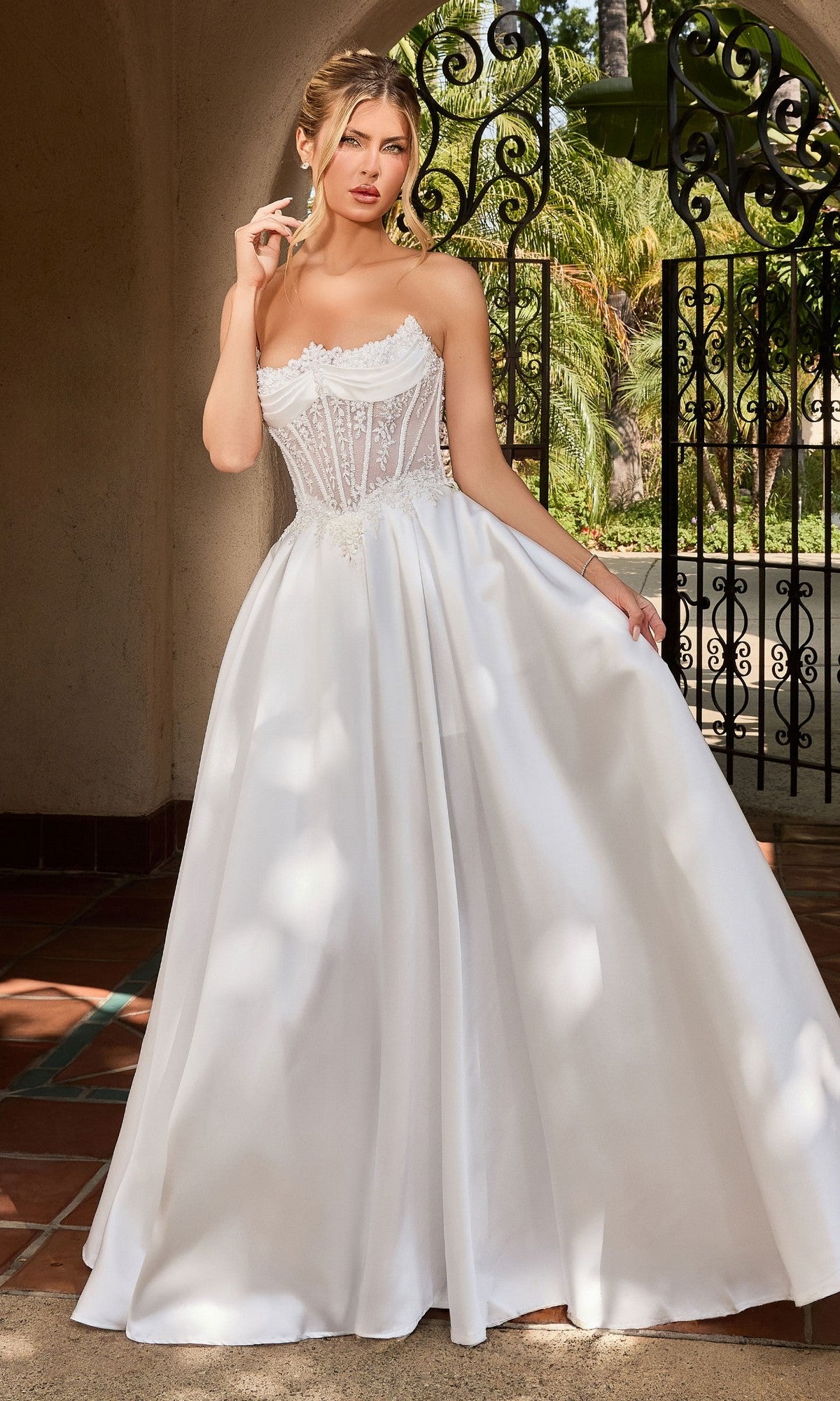 A woman in the Ladivine CC511W long formal dress in off white, featuring a strapless embroidered corset bodice and flowing A-line silhouette, stands outdoors before ornate wrought iron gates.
